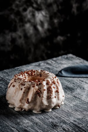 Photo for A delicious gugelhupf cake covered with a vanilla frosting plastic on a gray rustic wooden table, against a dark background with some blank space on top - Royalty Free Image