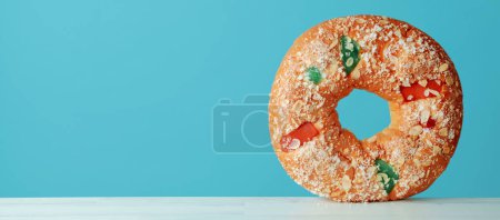 Téléchargez les photos : Un roscon de reyes, gâteau espagnol des trois rois mangé traditionnellement le jour de l'épiphanie, debout sur une table blanche devant un fond bleu, dans un format panoramique à utiliser comme bannière web ou en-tête - en image libre de droit