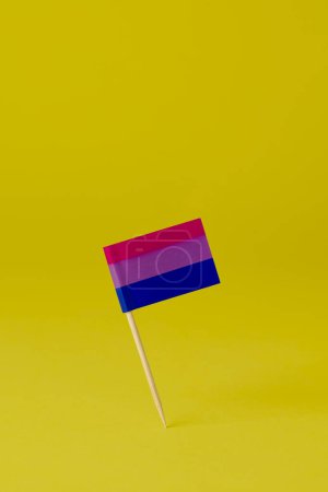 Foto de Una bandera de orgullo bisexual unida a un poste de madera se encuentra sobre un fondo amarillo, con un poco de espacio en blanco en la parte superior - Imagen libre de derechos