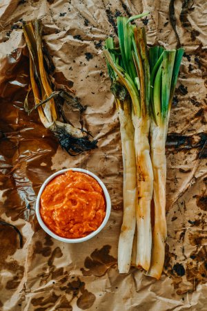 Photo for High angle view of some roasted calcots, the sweet onions typical of Catalonia, Spain, next to a ceramic bowl with some romesco sauce, on a crumpled kraft paper - Royalty Free Image