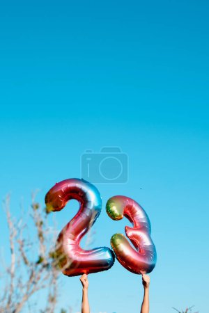 Foto de Un hombre sostiene dos globos multicolores en forma de número que forman el número 23 contra el cielo azul, con algo de espacio en blanco en la parte superior - Imagen libre de derechos