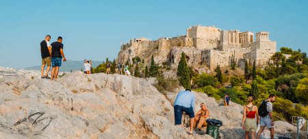 Foto de Atenas, Grecia - 29 de agosto de 2022: Algunas personas en la cima de una colina buscan el mejor lugar para tomar fotos de la Acrópolis de Atenas, en Grecia, en un formato panorámico - Imagen libre de derechos