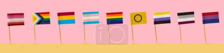 Téléchargez les photos : Certains drapeaux LGBTIQA différents attachés à des poteaux côte à côte sur une surface jaune, sur un fond rose, dans un format panoramique à utiliser comme bannière web - en image libre de droit