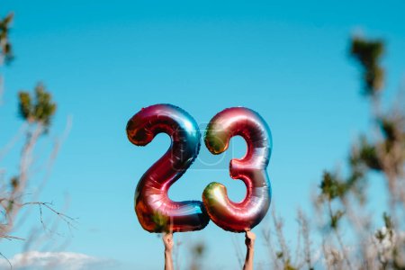 Foto de Primer plano de un hombre sosteniendo dos globos multicolores en forma de número que forman el número 23 en el cielo, de pie junto a algunas plantas y árboles al aire libre - Imagen libre de derechos