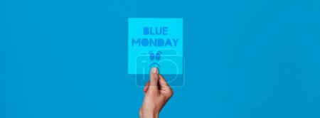 Téléchargez les photos : A man holds a blue sign with the text blue monday and a sad face against a blue background, in a panoramic format to use as web banner or header - en image libre de droit