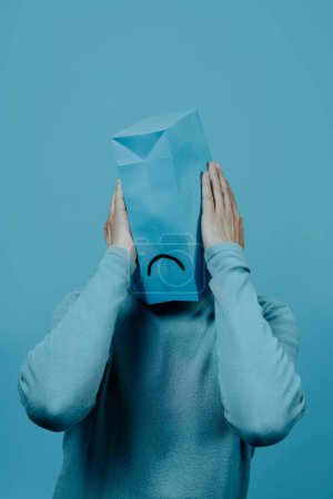 Photo for A man, wearing a blue paper bag in his head with a sad mouth drawn in it, has his hands around it, on a blue background - Royalty Free Image