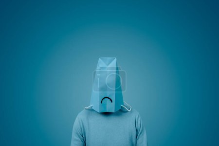 Photo for A man weas a blue paper bag in his head, with a sad mouth drawn in it, against a blue background with some blank space around him - Royalty Free Image