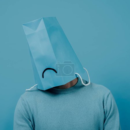 Téléchargez les photos : Closeup of a man wearing a blue paper bag in his head, with a sad mouth drawn in it, in front of a blue background - en image libre de droit