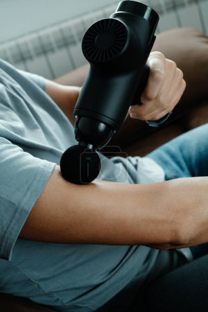 Téléchargez les photos : A caucasian man uses a massage gun to massage the muscles of his arm next to his elbow, while is sitting on a couch - en image libre de droit