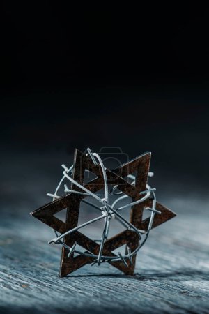 Téléchargez les photos : An old star of david tied with some barbed wire on a gray rustic wooden surface, against a black background with some blank space on top - en image libre de droit