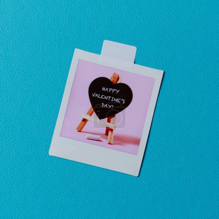 Photo for Closeup of a retro instant photo, that shows a heart-shaped blackboard with text happy valentines day written in it, attached to a  blue leatherette surface - Royalty Free Image