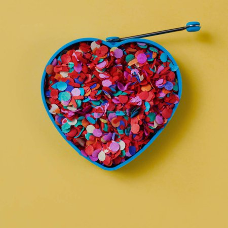 Photo for High angle view of a blue heart-shaped mould full of confetti of different colors, on a yellow background, in a square format - Royalty Free Image
