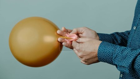 Foto de Un hombre utiliza una herramienta para atar un globo dorado inflado, sobre un fondo gris, en un formato panorámico para usar como banner web o encabezado - Imagen libre de derechos