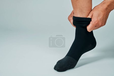 Téléchargez les photos : A man puts on a black compression sock in front of an off-white background with some blank space on the left - en image libre de droit