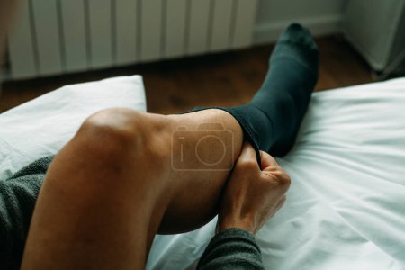 Foto de A man is putting on a compression sock sitting on his bed at home - Imagen libre de derechos