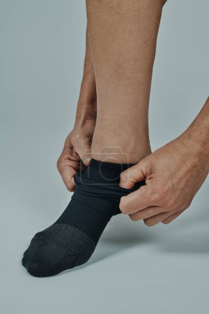 Téléchargez les photos : Un homme est sur le point de mettre une chaussette de compression devant un fond blanc cassé - en image libre de droit
