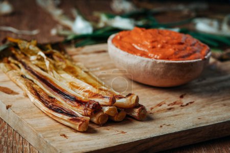 Téléchargez les photos : Some roasted calcots, the sweet onions typical of Catalonia, Spain, and a rustic ceramic bowl with some romesco sauce on a wooden cutting board - en image libre de droit
