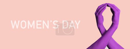 Téléchargez les photos : Une paire de mains, peintes en violet, formant un ruban de sensibilisation violet et teh texte journée des femmes sur un fond rose, dans un format panoramique à utiliser comme bannière web - en image libre de droit
