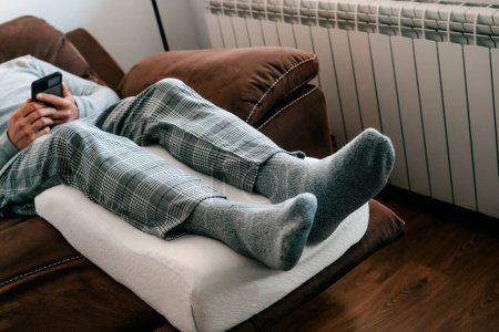 Téléchargez les photos : Un homme appuie ses jambes sur un oreiller d'élévation de jambe, fait de mousse de mémoire, tout en se reposant sur le canapé - en image libre de droit