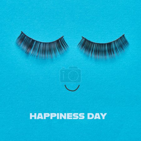 Foto de A pair of fake eyelashes and a smiling mouth forming a happy face, and the text happiness day on a blue background - Imagen libre de derechos