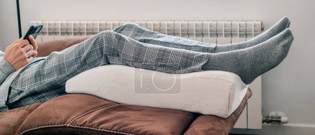 Foto de Un hombre apoya sus piernas en una almohada de elevación de piernas hecha de espuma de memoria, descansando en el sofá, en un formato panorámico para usar como banner web o encabezado - Imagen libre de derechos