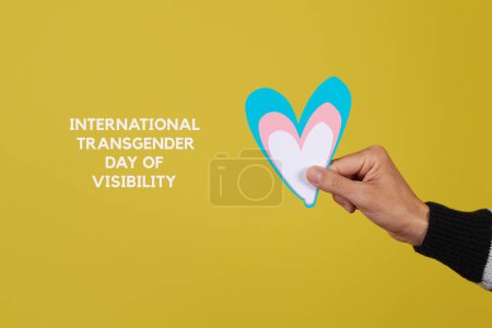 Téléchargez les photos : La main d'un jeune caucasien tenant un cœur aux couleurs du drapeau transgenre et le texte la journée internationale de visibilité transgenre sur fond jaune - en image libre de droit