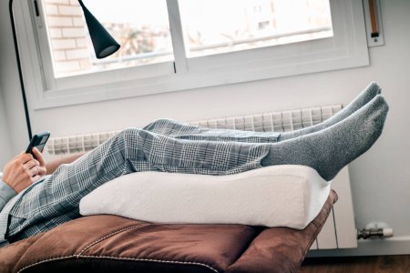 Foto de Primer plano de un hombre descansando en el sofá apoyando sus piernas en una almohada de elevación de la pierna, hecha de espuma de memoria - Imagen libre de derechos