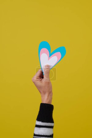 Foto de Una joven caucásica sostiene un corazón con los colores de la bandera transgénero frente a un fondo amarillo con un poco de espacio en blanco en la parte superior - Imagen libre de derechos