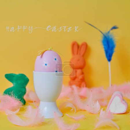 Téléchargez les photos : Texte joyeux Pâques et un oeuf de Pâques rose dans un oeuf blanc, quelques plumes roses et quelques lapins de Pâques de différentes couleurs sur un fond jaune - en image libre de droit
