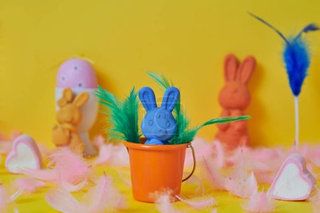 Photo for A blue easter bunny in an orange bucket surrounded by pink feathers and a pink easter egg and some other easter bunnies of different colors on a yellow background - Royalty Free Image
