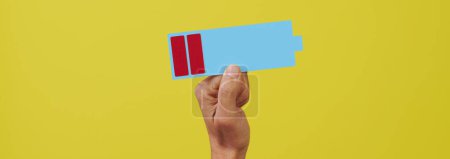 Téléchargez les photos : Un homme tenant une représentation en carton d'une batterie basse sur un fond jaune, dans un format panoramique à utiliser comme bannière Web ou en-tête - en image libre de droit