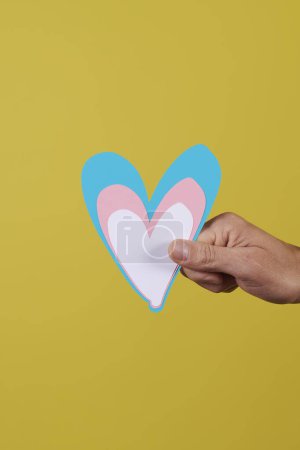 Foto de Primer plano de la mano de un joven caucásico sosteniendo un corazón con los colores de la bandera transgénero sobre un fondo amarillo - Imagen libre de derechos