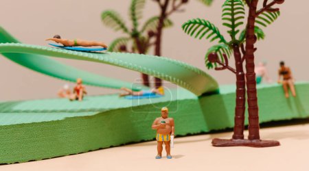 Téléchargez les photos : Certaines personnes miniatures, en maillot de bain, sur et autour d'un flip-flop vert, à côté d'un palmier miniature, sur un fond brun pâle - en image libre de droit