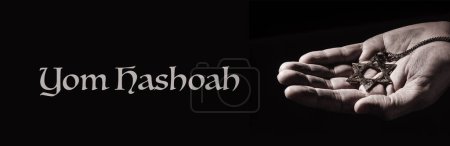 Téléchargez les photos : La main d'un homme avec un pendentif vieux et rouillé en forme d'étoile de David et le texte Yom Hashoah sur fond noir, dans un format panoramique à utiliser comme bannière web - en image libre de droit