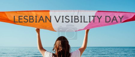 Téléchargez les photos : Le texte journée de visibilité lesbienne et une femme tenant un drapeau de fierté lesbienne au-dessus de sa tête sur le ciel, dans un format panoramique à utiliser comme bannière web - en image libre de droit
