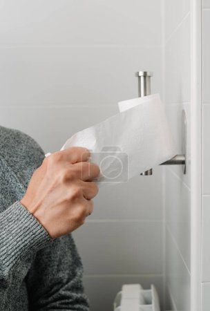 Foto de Primer plano de un hombre, sentado en el inodoro, tomando un pedazo de papel higiénico, del rollo de papel colocado en el portarrollos - Imagen libre de derechos