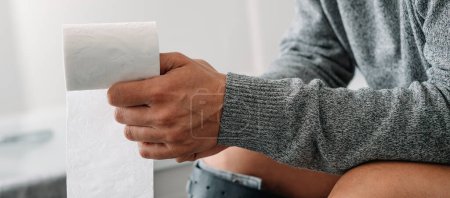 Foto de Un hombre caucásico, sentado en el inodoro, tiene un rollo de papel higiénico en la mano, en un formato panorámico para usar como banner web o encabezado - Imagen libre de derechos