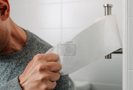 Téléchargez les photos : Un homme, assis dans les toilettes, prend un morceau de papier toilette, du rouleau de papier placé dans le porte-rouleau de toilette - en image libre de droit