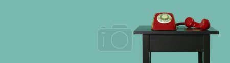 Téléchargez les photos : Un téléphone à cadran rotatif fixe rouge, avec son combiné décroché sur une table en bois noir, sur un fond bleu dans un format panoramique à utiliser comme bannière Web ou en-tête - en image libre de droit