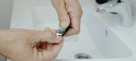 Foto de Un hombre se corta las uñas con un cortaúñas, apoyando su mano en el lavabo blanco de su baño, en un formato panorámico para usar como banner web - Imagen libre de derechos