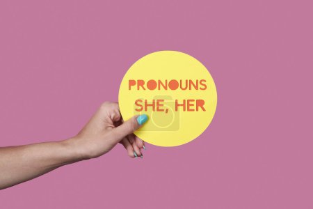 Foto de Primer plano de una persona caucásica sosteniendo un signo amarillo con el texto mis pronombres son ella, ella sobre un fondo rosa - Imagen libre de derechos