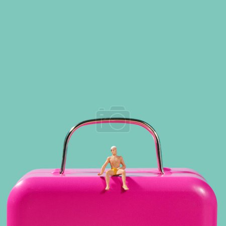 Photo for A miniature man wearing swimsuit sitting on top of a pink suitcase against a blue background with some blank space on top - Royalty Free Image