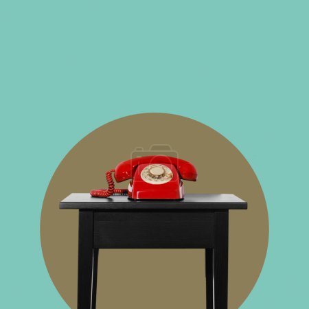 Foto de Un teléfono de marcación rotatoria fijo rojo en una mesa negra frente a un círculo verde oliva y un fondo azul con un poco de espacio en blanco en la parte superior - Imagen libre de derechos