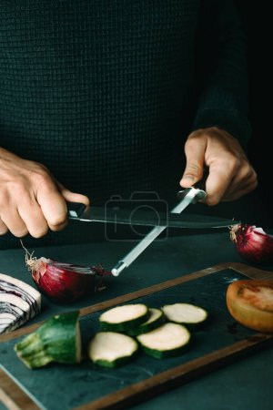 Photo for Man steeling a kitchen knife with a honing steel at a table with some chopped vegetables - Royalty Free Image