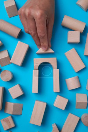 Photo for High angle view of a man building a house with some wooden building blocks on a blue background surrounded by some more building blocks - Royalty Free Image