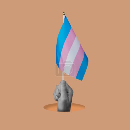 Foto de La mano de una persona en blanco y negro ondeando una bandera de orgullo transgénero asomándose por un agujero sobre un fondo rosa anaranjado - Imagen libre de derechos