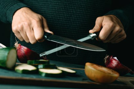 Photo for A man steels a kitchen knife with a honing steel at a table with some vegetables - Royalty Free Image