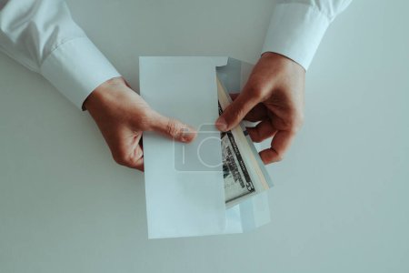 Photo for High angle view of a man wearing a white long-sleeve shirt putting, or taking out, a bunch of dollar notes in an envelope, sitting at a white table - Royalty Free Image