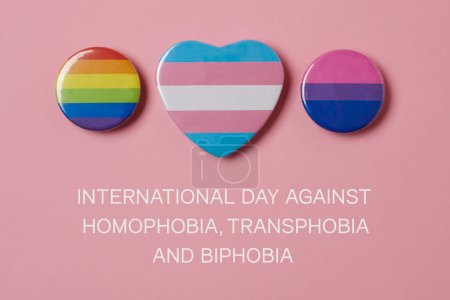Foto de Algunas insignias con una bandera de orgullo gay, una bandera de orgullo transgénero y una bandera de orgullo bisexual, y el texto del día internacional contra la homofobia, la transfobia y la bifobia en un fondo rosa - Imagen libre de derechos