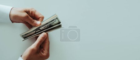 Hochwinkelaufnahme eines Mannes in einem weißen Langarmhemd, der an einem weißen Tisch ein paar Dollarnoten zählt, im Panoramaformat, um sie als Web-Banner zu verwenden